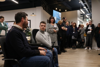 Two men, one in a wheelchair, discussing accessibility in front of room of people