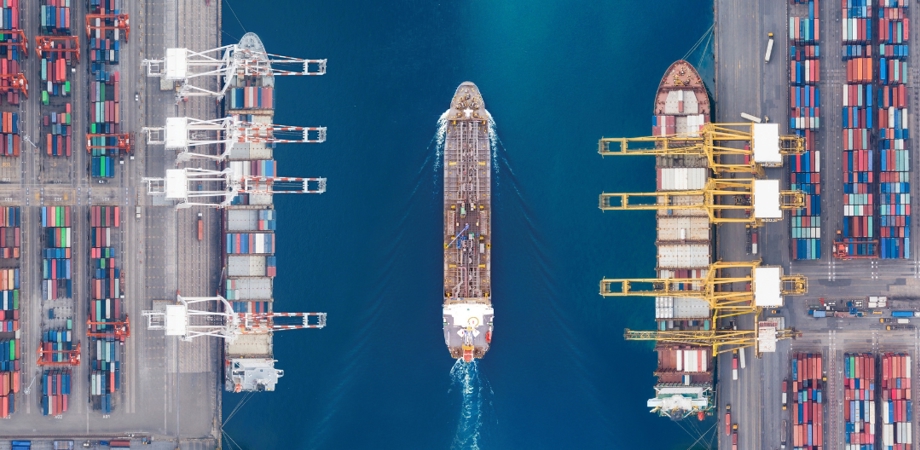 An overhead image of a container ship on a bright blue ocean sailing between two cranes at port