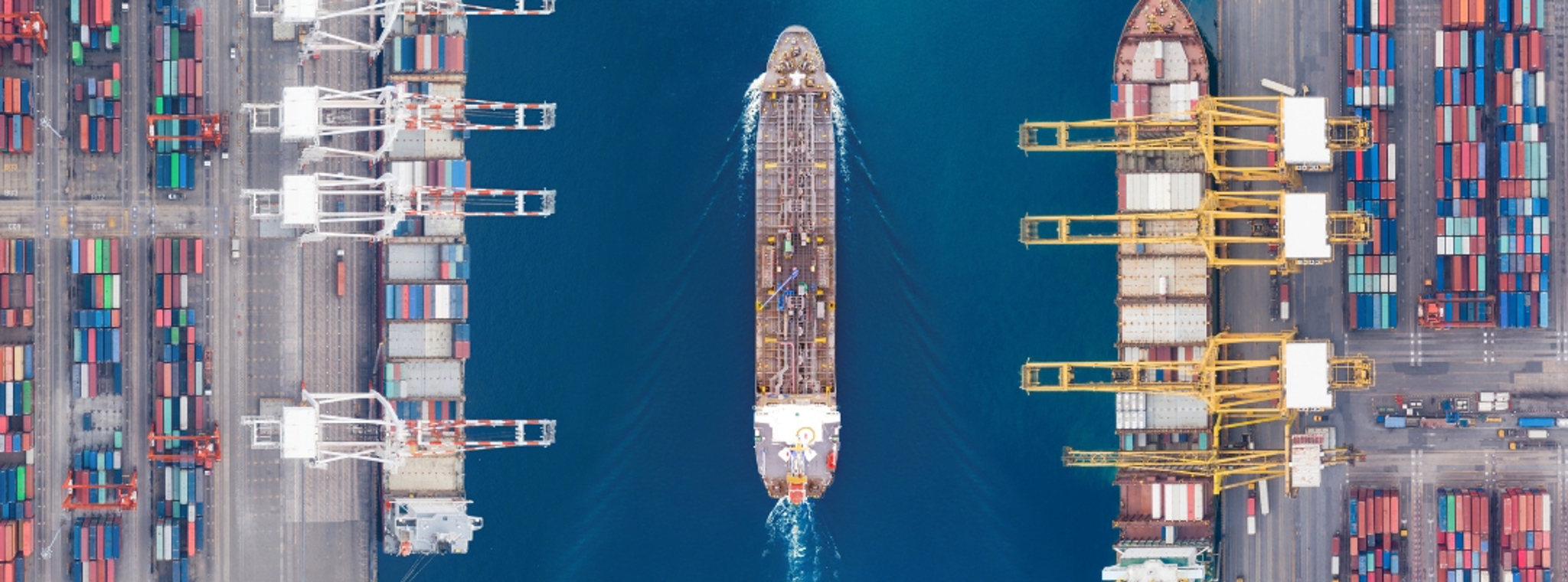 An overhead image of a container ship on a bright blue ocean sailing between two cranes at port
