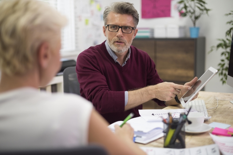 An image of a senior worker at a desk talking to a colleague while referring to an iPad
