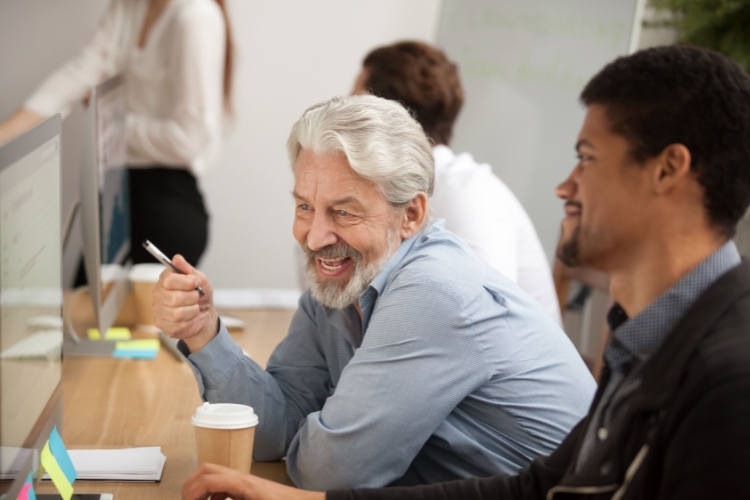 Senior man with a coffee cup sat next to a young man working on computer