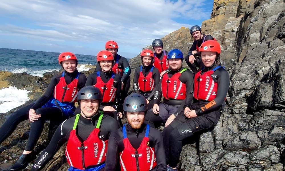 A group of people wearing coasteering outfits sat on cliff face